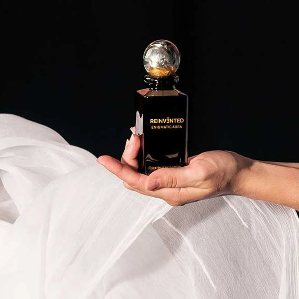 Hand holding a black perfume bottle labeled 'Reinvented Enigmatic Aura' against a dark background.