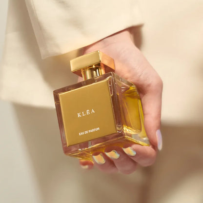 Hand holding a gold 'KLEA' perfume bottle against a neutral background
