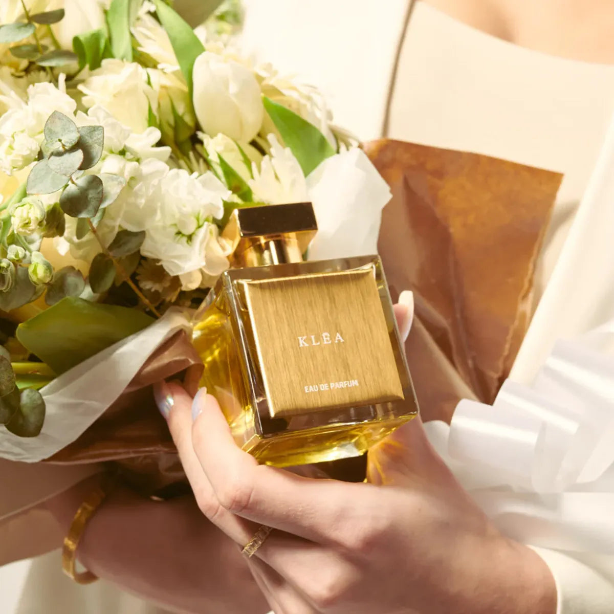 Gold perfume bottle with 'KLEA' branding held by a person next to a bouquet of flowers.